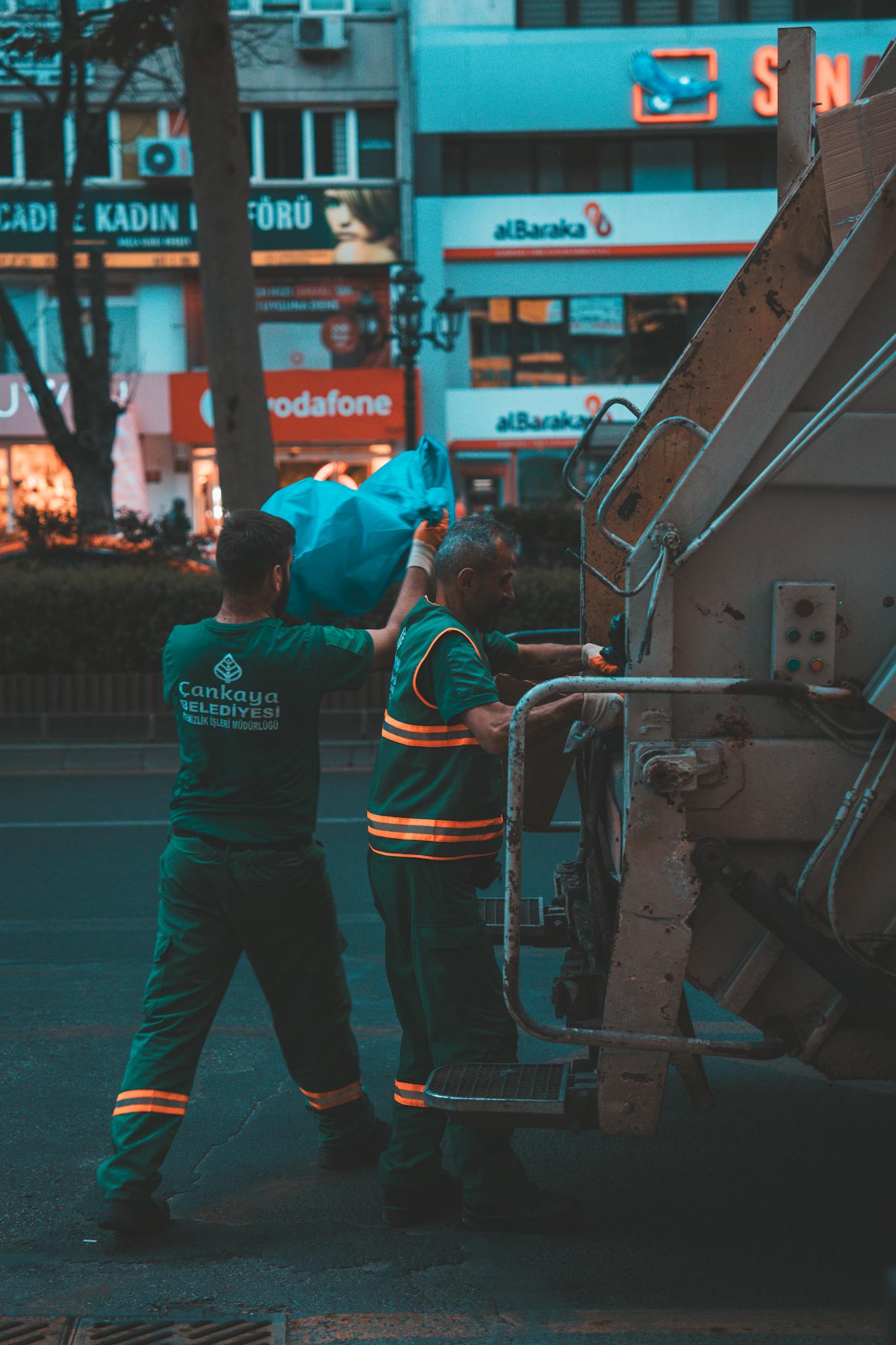 Two city workers collect garbage in an urban setting during early morning hours.